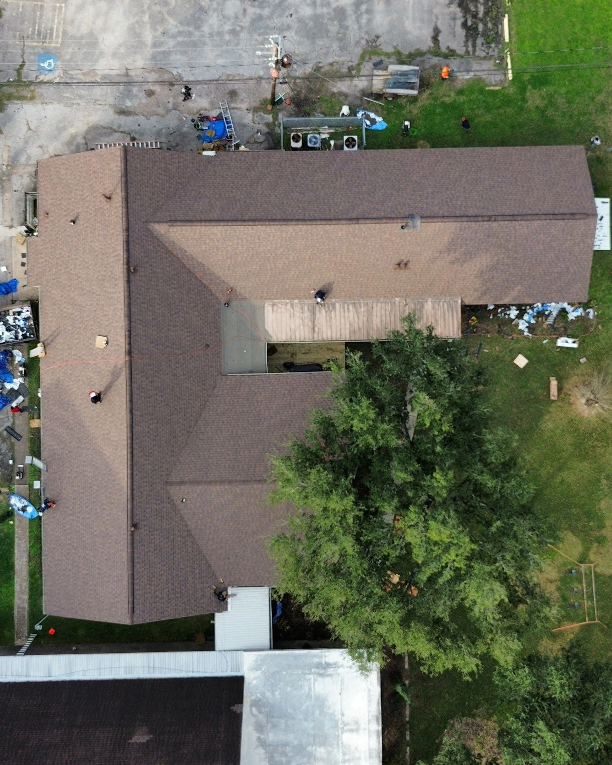 Aerial view of a large building with a newly installed brown shingle roof, showing clean rooflines and surrounding parking area, grass, and equipment used for the roofing project.